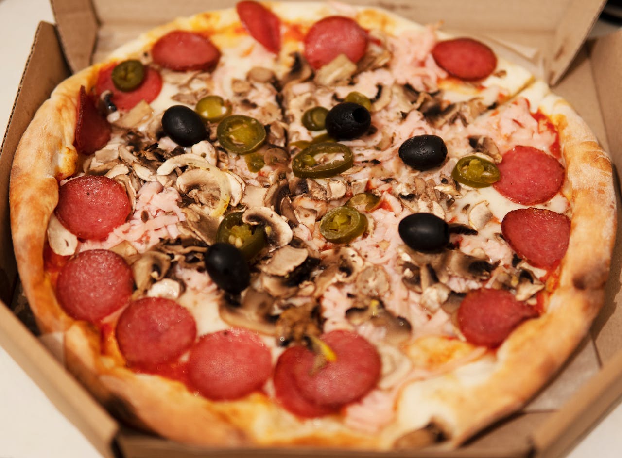 Close-up of a freshly baked pizza topped with pepperoni, mushrooms, olives, and jalapenos in a cardboard box.